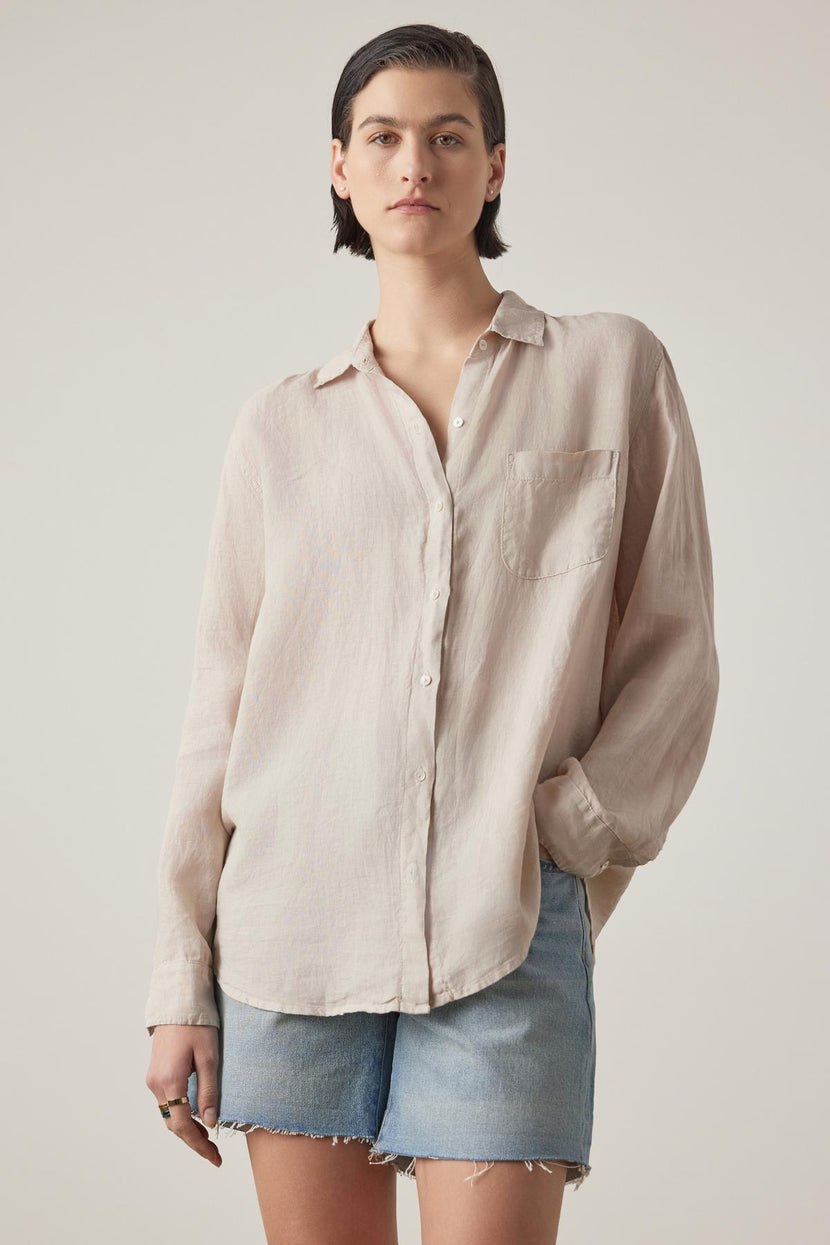 Person with short dark hair wearing a Velvet by Jenny Graham MULHOLLAND LINEN SHIRT featuring a relaxed silhouette and scooped hemline, paired with light blue denim shorts, stands against a plain background.
