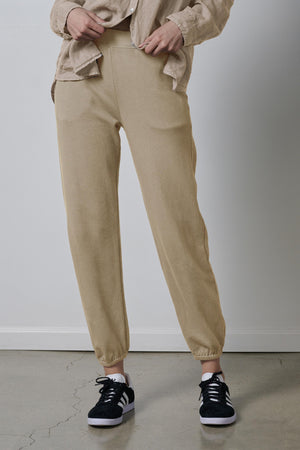 A person standing in a neutral pose wearing Velvet by Jenny Graham's ZUMA SWEATPANT, a beige jacket, and black sneakers.