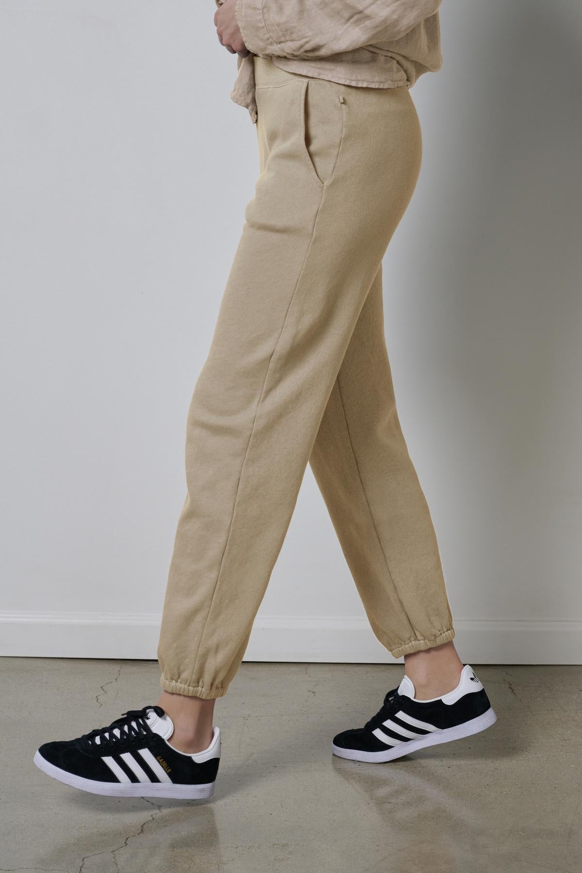 Person wearing sustainable fashion, Velvet by Jenny Graham's ZUMA SWEATPANT beige organic cotton joggers and black sneakers with white stripes.-36463615672513
