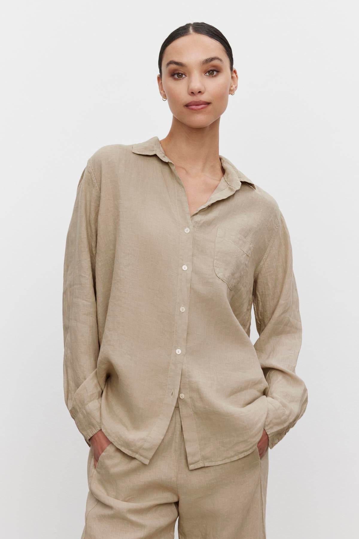 A person wearing the MULHOLLAND LINEN SHIRT in light beige by Velvet by Jenny Graham, paired with matching pants, stands against a plain white background. Their hands are in their pockets, and they have a slight smile, exuding a relaxed silhouette.-37618568069313