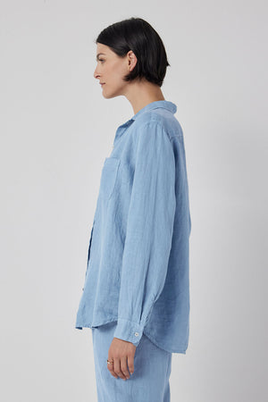 A woman with a relaxed silhouette is seen from the back wearing a blue linen Mulholland shirt by Velvet by Jenny Graham with a scooped hemline.