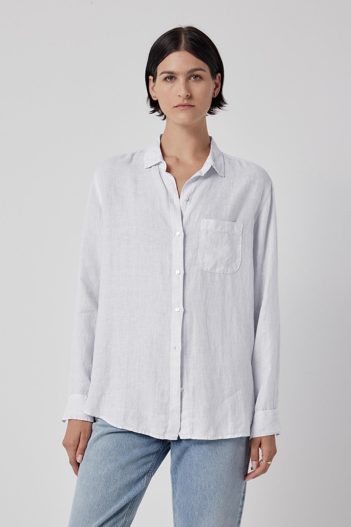 A woman wearing a Velvet by Jenny Graham Mulholland shirt in casual white linen with a scooped hemline and blue jeans stands against a grey background.-36463556231361