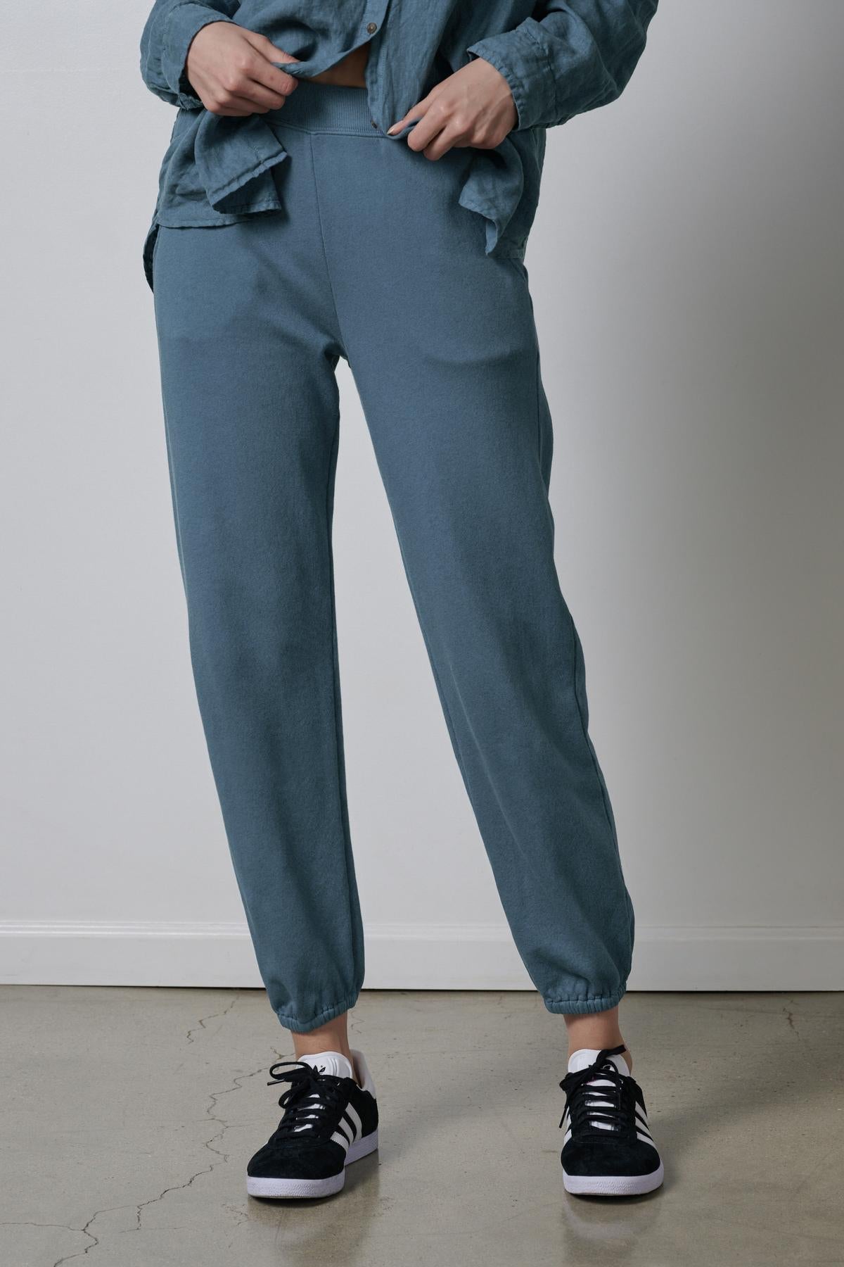 A person standing against a neutral background, wearing a blue button-up shirt and matching blue Velvet by Jenny Graham ZUMA SWEATPANTS with black sneakers.-36594728009921