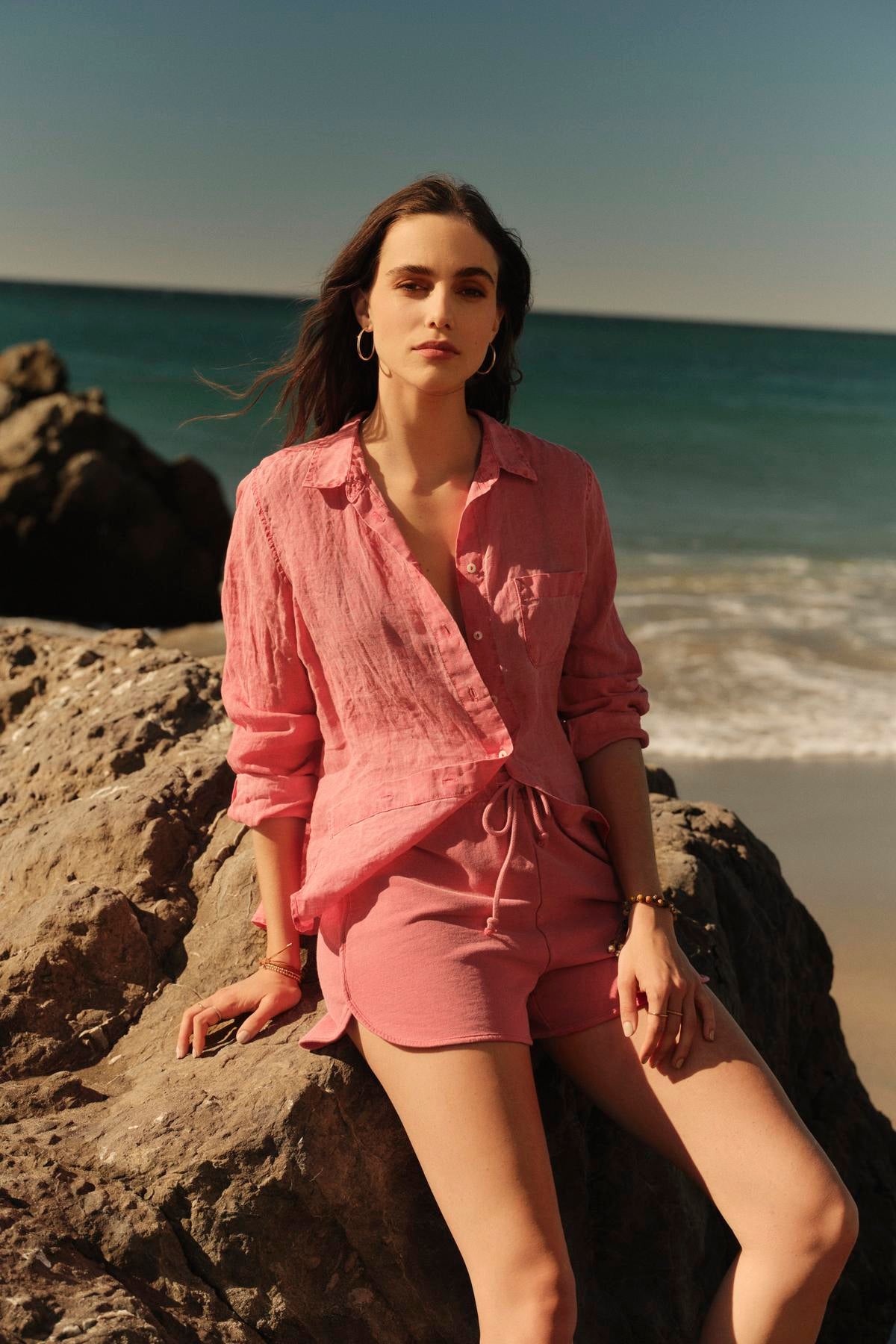   A person in a relaxed fit pink shirt and PRESELY SHORT by Velvet by Graham & Spencer sits on a rock by the beach, with the ocean and sky in the background. 