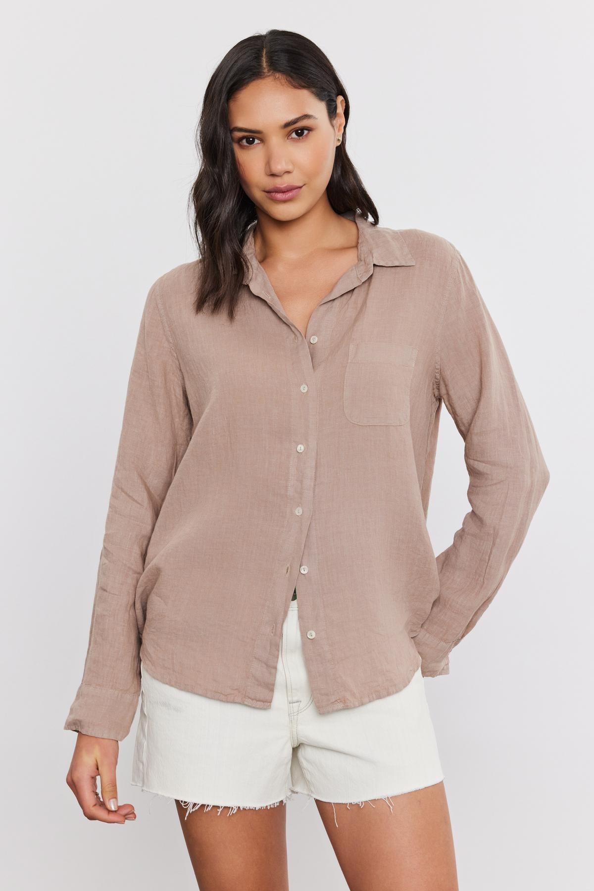   A woman with shoulder-length dark hair wearing a casual beige Velvet by Graham & Spencer Natalia Linen Button-Up Shirt and white frayed shorts, both in a relaxed silhouette. 