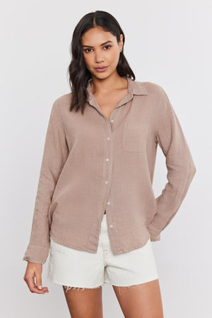 A woman with shoulder-length dark hair wearing a casual beige Velvet by Graham & Spencer Natalia Linen Button-Up Shirt and white frayed shorts, both in a relaxed silhouette.