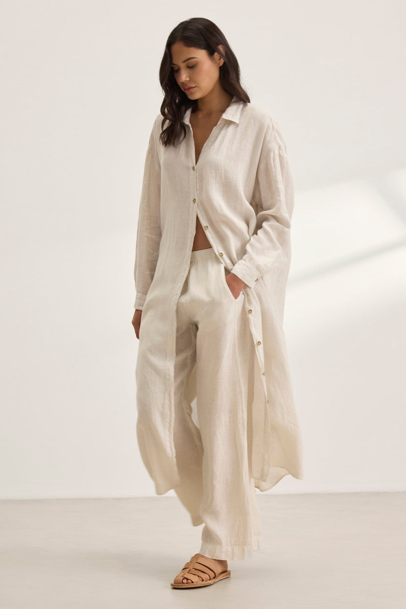 A woman wearing the Velvet by Graham & Spencer NICOLE LINEN DRESS and matching wide-leg pants stands in a neutral room with soft shadows, embodying breathable comfort.