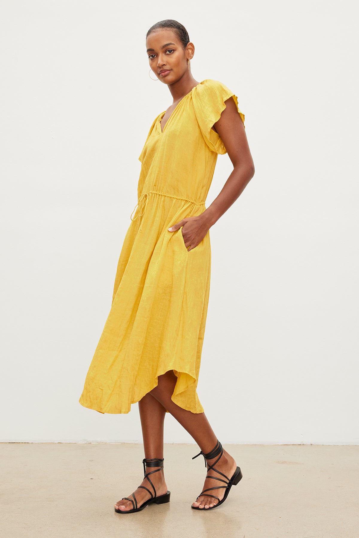   A woman in a yellow Velvet by Graham & Spencer PEPPER LINEN V-NECK DRESS with a drawstring waist and black sandals stands confidently, her hands placed slightly on her hips, posing against a white background. 