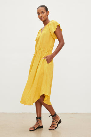 A woman in a yellow Velvet by Graham & Spencer PEPPER LINEN V-NECK DRESS with a drawstring waist and black sandals stands confidently, her hands placed slightly on her hips, posing against a white background.