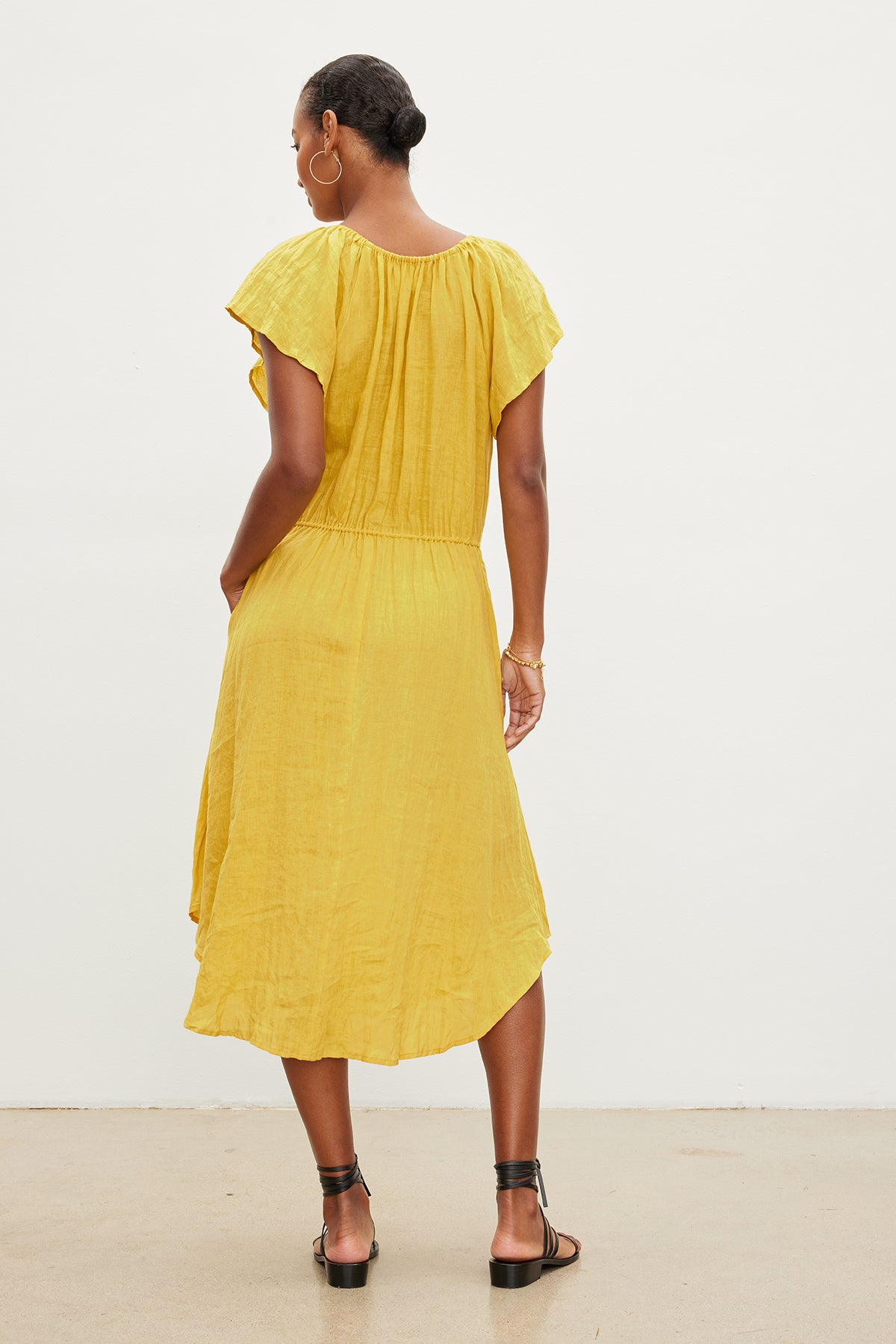 Woman seen from behind wearing a yellow Velvet by Graham & Spencer PEPPER LINEN V-NECK DRESS with ruffled sleeves and pleated back, paired with black heeled sandals.-35967692013761