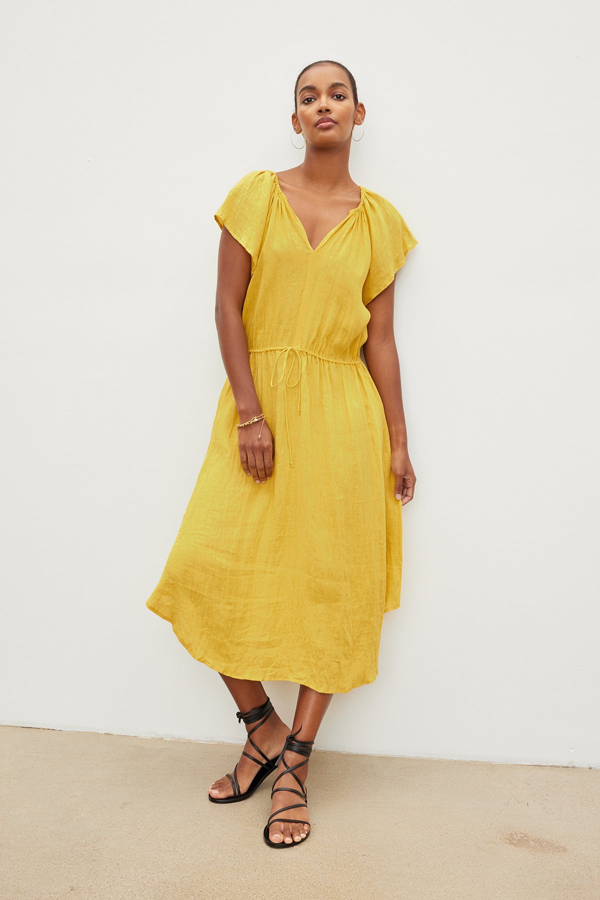   Woman in a yellow PEPPER LINEN V-NECK DRESS by Velvet by Graham & Spencer with a drawstring waist and black sandals stands against a white wall, looking directly at the camera. 