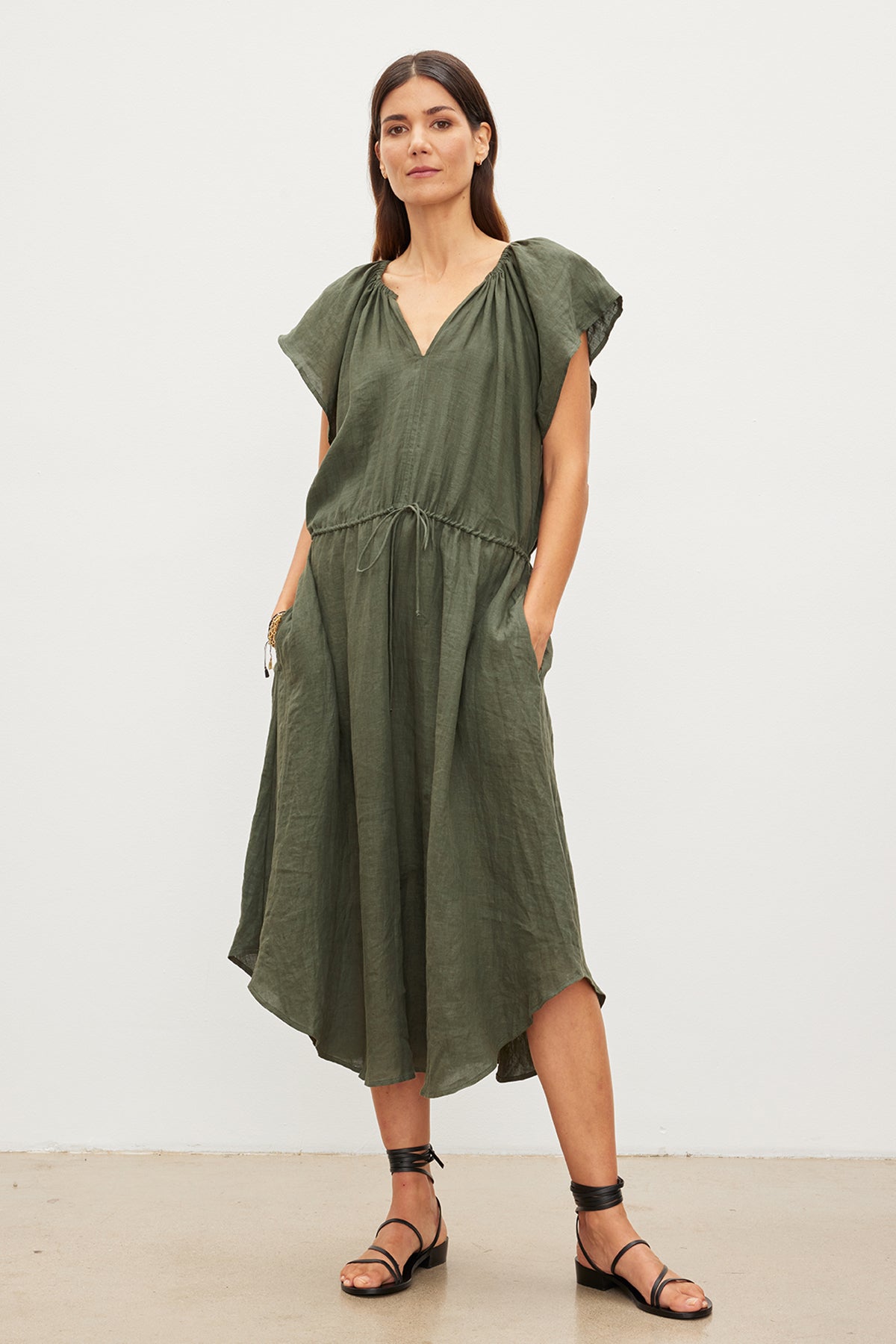   A woman stands in a studio, wearing a Velvet by Graham & Spencer green woven linen midi dress with a drawstring waist and black sandals. She has shoulder-length brown hair and a neutral expression. 