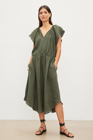 A woman stands in a studio, wearing a Velvet by Graham & Spencer green woven linen midi dress with a drawstring waist and black sandals. She has shoulder-length brown hair and a neutral expression.