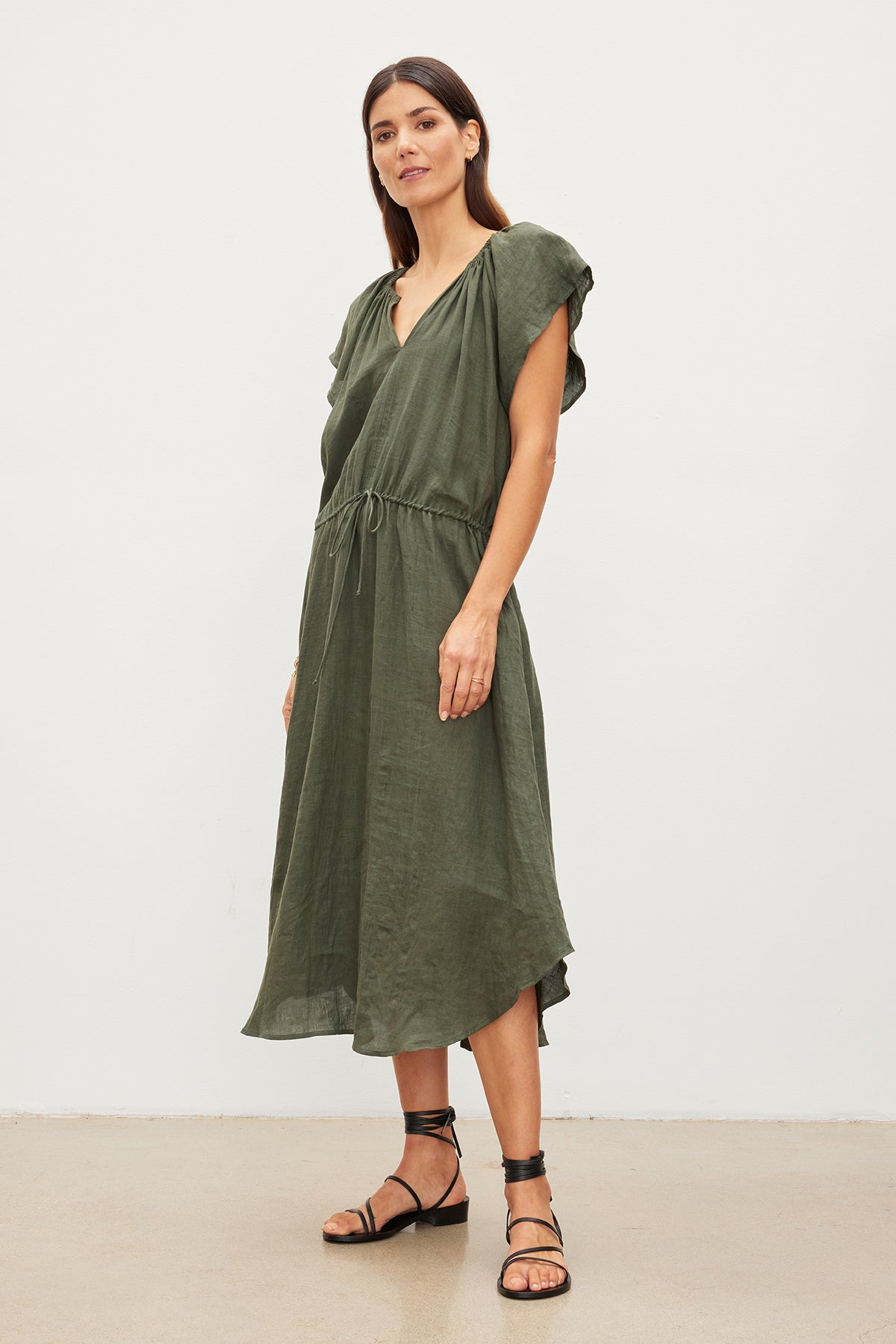 A woman in a Velvet by Graham & Spencer Pepper Linen V-Neck Dress with a v-neckline and ruffled sleeves, paired with black strappy sandals, standing in a studio setting.-35967692144833
