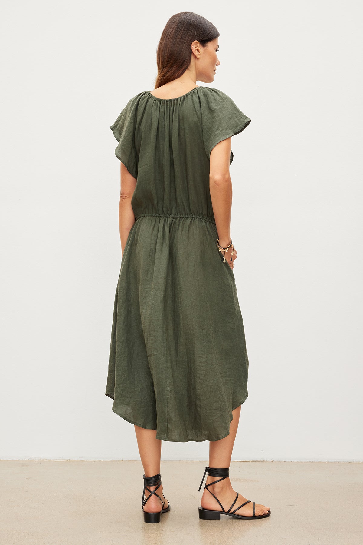 A woman stands facing away from the camera, wearing a green woven Pepper Linen V-Neck Dress with ruffled sleeves and black sandals by Velvet by Graham & Spencer.-35967692177601