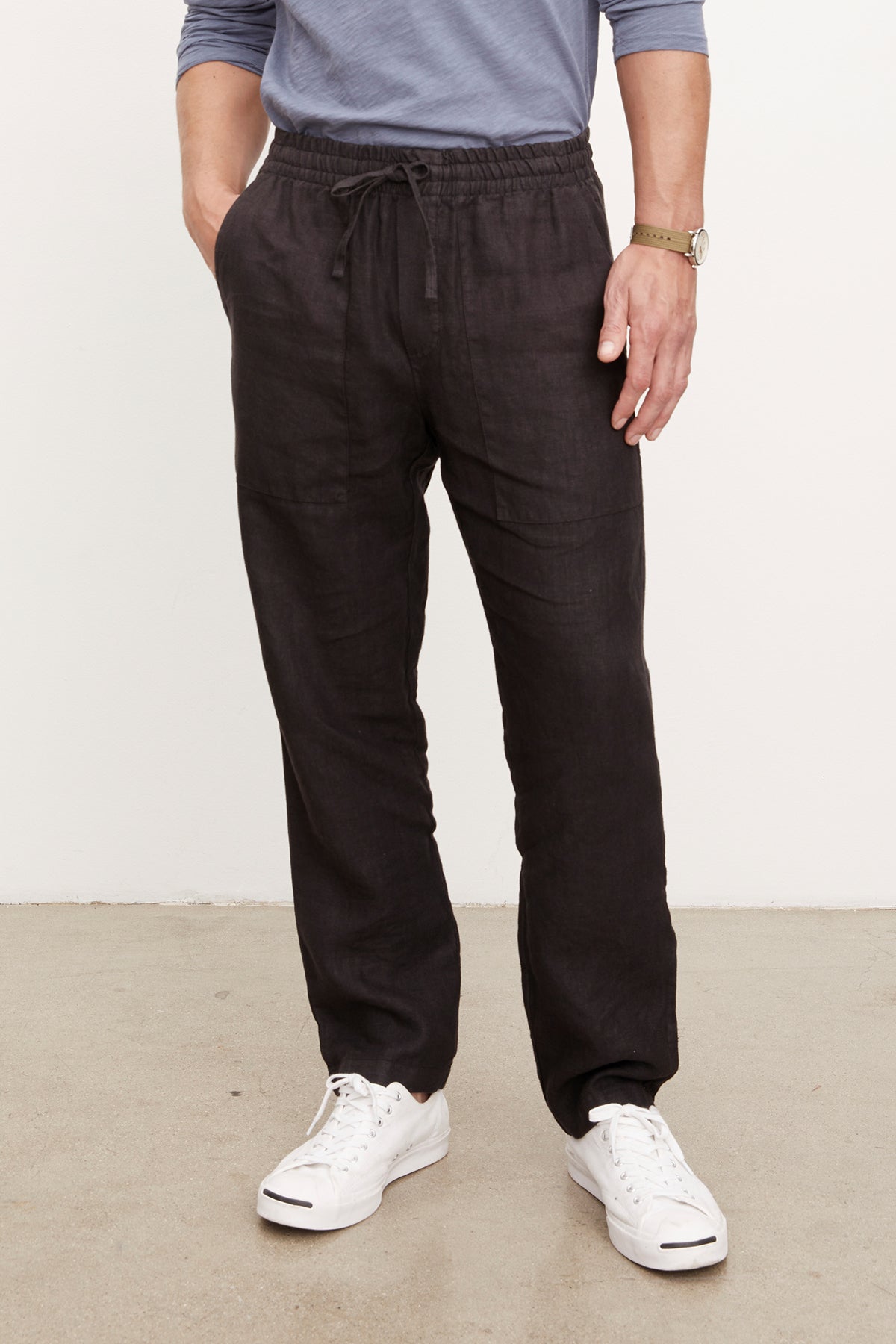   Wearing Velvet by Graham & Spencer's PHELAN LINEN PANT in black, a person with relaxed fit, elastic drawstring waist pants stands against a light background, paired with a blue long-sleeve shirt, white sneakers, and a wristwatch. 