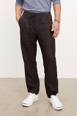 Wearing Velvet by Graham & Spencer's PHELAN LINEN PANT in black, a person with relaxed fit, elastic drawstring waist pants stands against a light background, paired with a blue long-sleeve shirt, white sneakers, and a wristwatch.