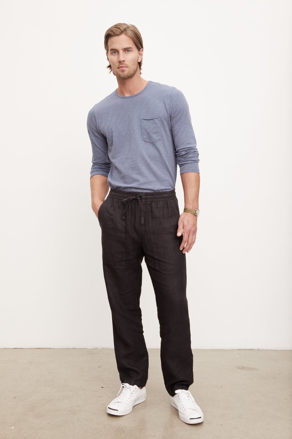  A man in a blue t-shirt and jeans is standing in front of a white wall wearing Velvet by Graham & Spencer's Phelan Linen Pant. 