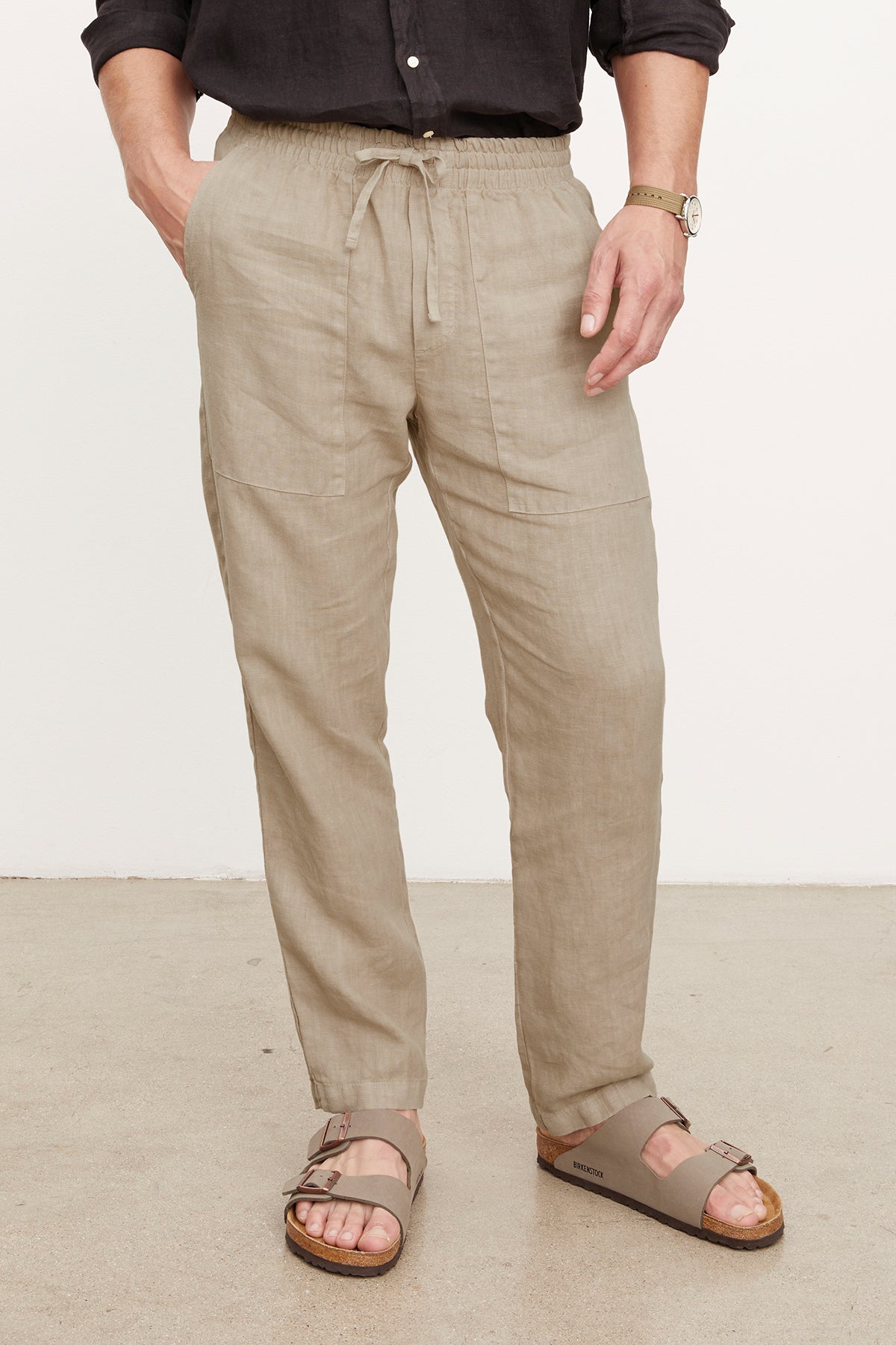   Wearing Velvet by Graham & Spencer's PHELAN LINEN PANT with a relaxed fit and elastic drawstring waist, a person pairs them with a dark shirt, watch, and taupe sandals while standing on a light-colored floor. 