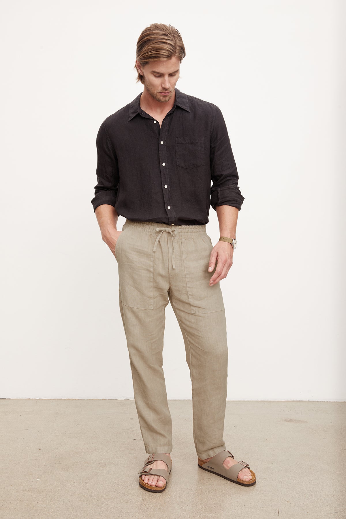   A man stands in a studio, dressed in a dark shirt tucked into Velvet by Graham & Spencer Phelan Linen Pants and sandals, gazing downwards with hands in pockets. 