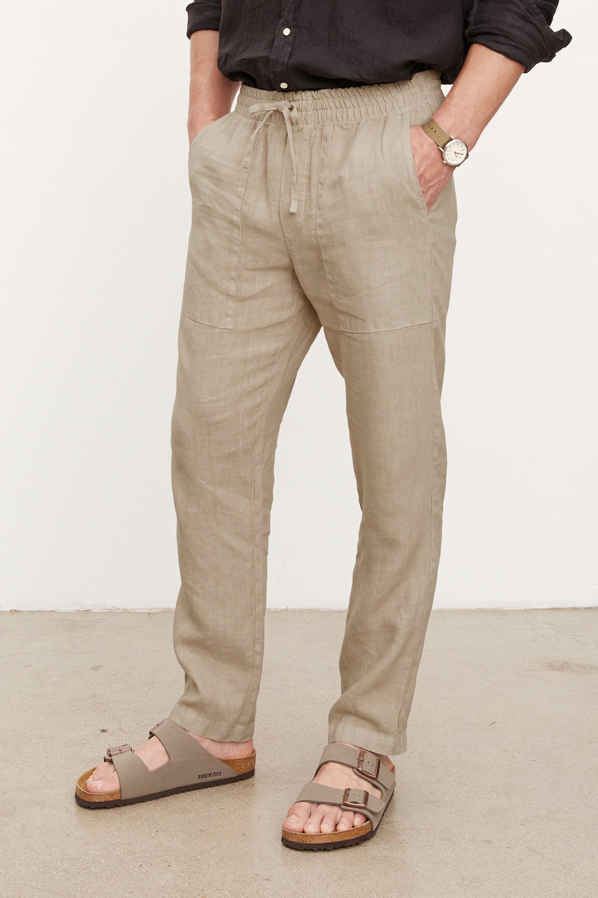   Man standing in a casual pose wearing Velvet by Graham & Spencer's Phelan Linen Pants, a black shirt, a watch, and beige sandals. Only the lower half of his body is visible. 