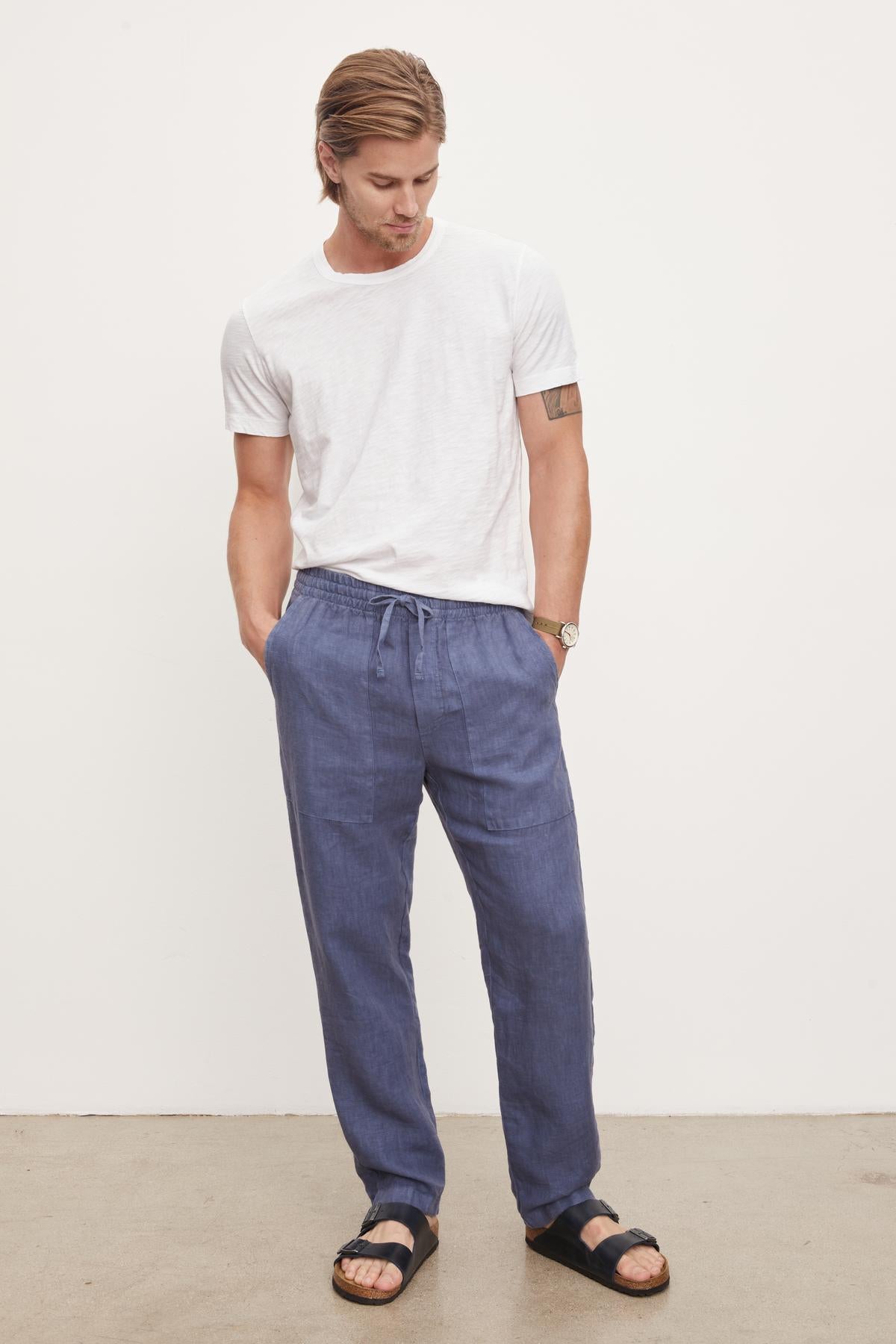   A man wearing a white t-shirt, Velvet by Graham & Spencer blue Phelan linen pants, and black sandals stands looking down, against a plain background. 