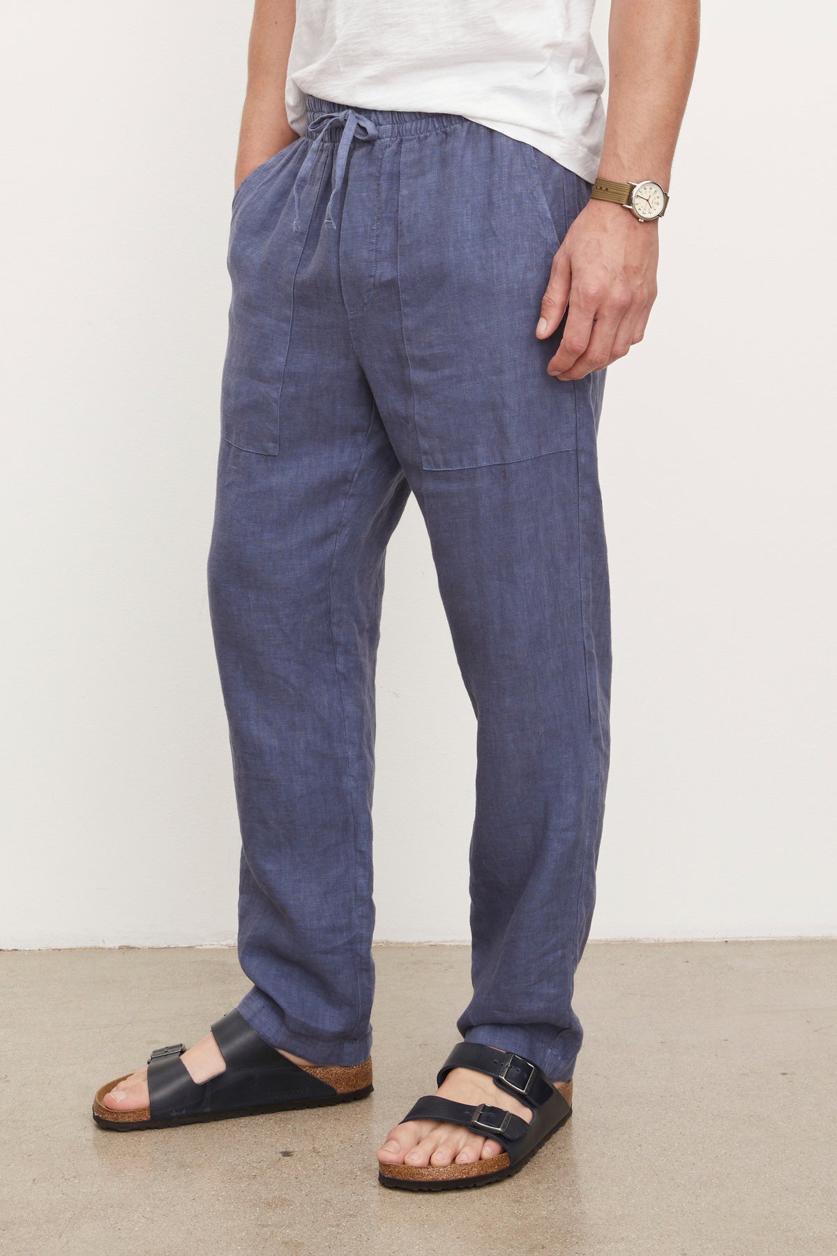 A person wearing blue Velvet by Graham & Spencer Phelan Linen Pants, black sandals, and a silver wristwatch, standing against a plain wall. Visible from the waist down.-36805503418561