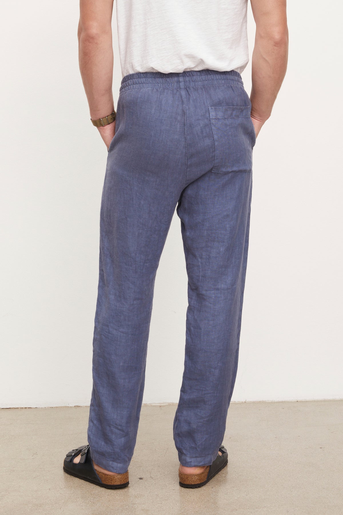   Rear view of a person in a white top and blue relaxed fit Velvet by Graham & Spencer Phelan Linen Pant, standing in a room with white walls. 