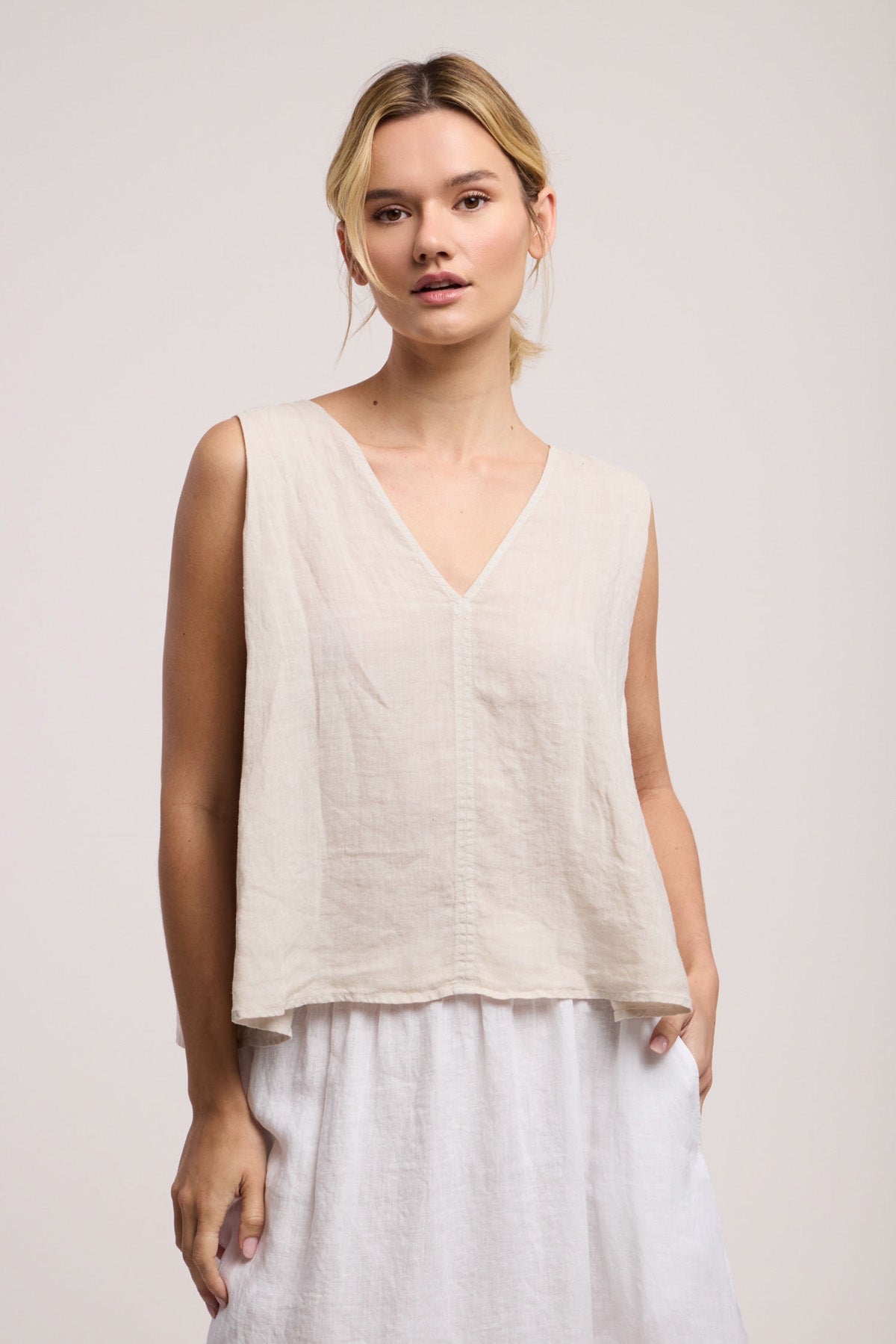 A woman wears the RENNA LINEN TOP by Velvet by Graham & Spencer, a sleeveless, light beige V-neck summer essential, paired with a white skirt against a plain background.-41371968274625