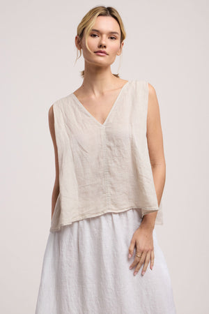 A person wears the RENNA LINEN TOP by Velvet by Graham & Spencer—a sleeveless V-neck paired with a white skirt—standing against a plain light background, capturing the essence of a summer essential.