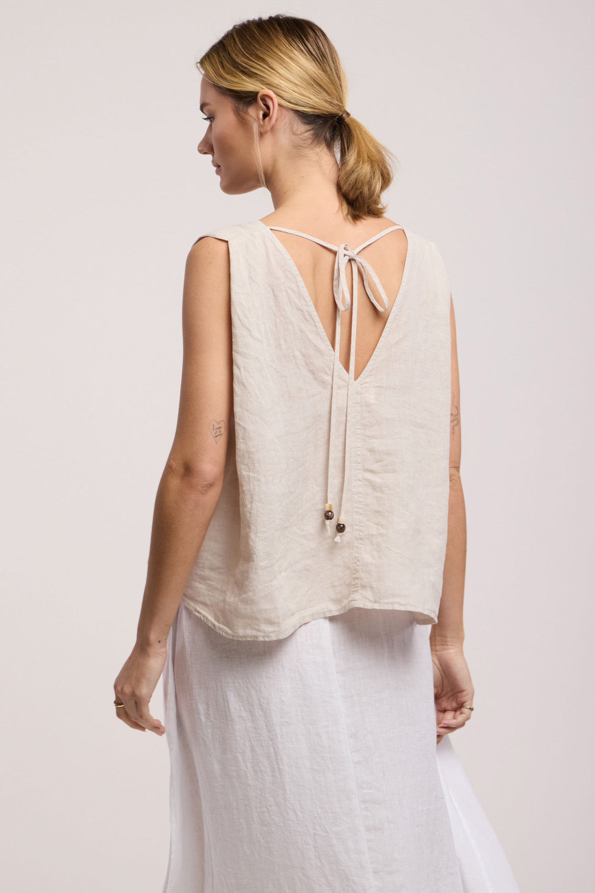 A woman wears the RENNA LINEN TOP by Velvet by Graham & Spencer, a sleeveless summer essential with a back tie, paired with a white skirt against a neutral background.