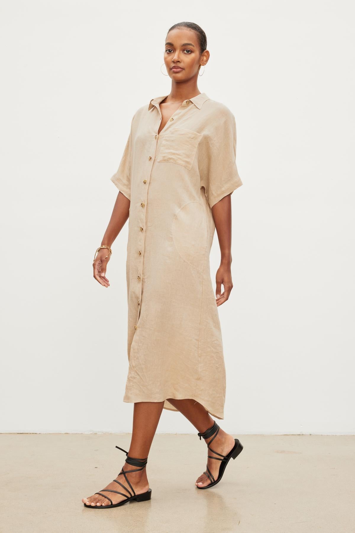   A woman in a SANDRA LINEN DRESS by Velvet by Graham & Spencer, complete with a detachable belt, and black sandals walks across a plain background. 