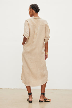 A person stands against a plain background, wearing the light brown SANDRA LINEN DRESS with rolled-up sleeves by Velvet by Graham & Spencer, black strappy sandals, and a detachable belt. They are facing away from the camera.