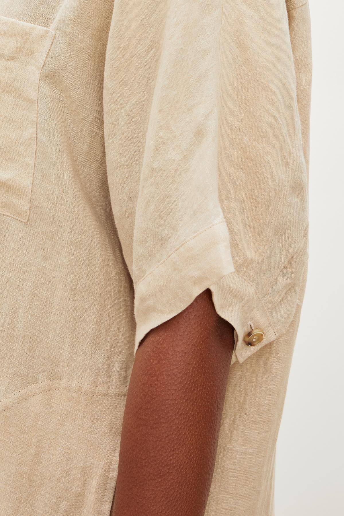   Close-up of a person wearing the SANDRA LINEN DRESS by Velvet by Graham & Spencer, showcasing its beige color, short sleeves with a button-up design, and visible chest pocket. 