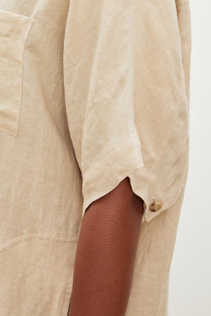 Close-up of a person wearing the SANDRA LINEN DRESS by Velvet by Graham & Spencer, showcasing its beige color, short sleeves with a button-up design, and visible chest pocket.