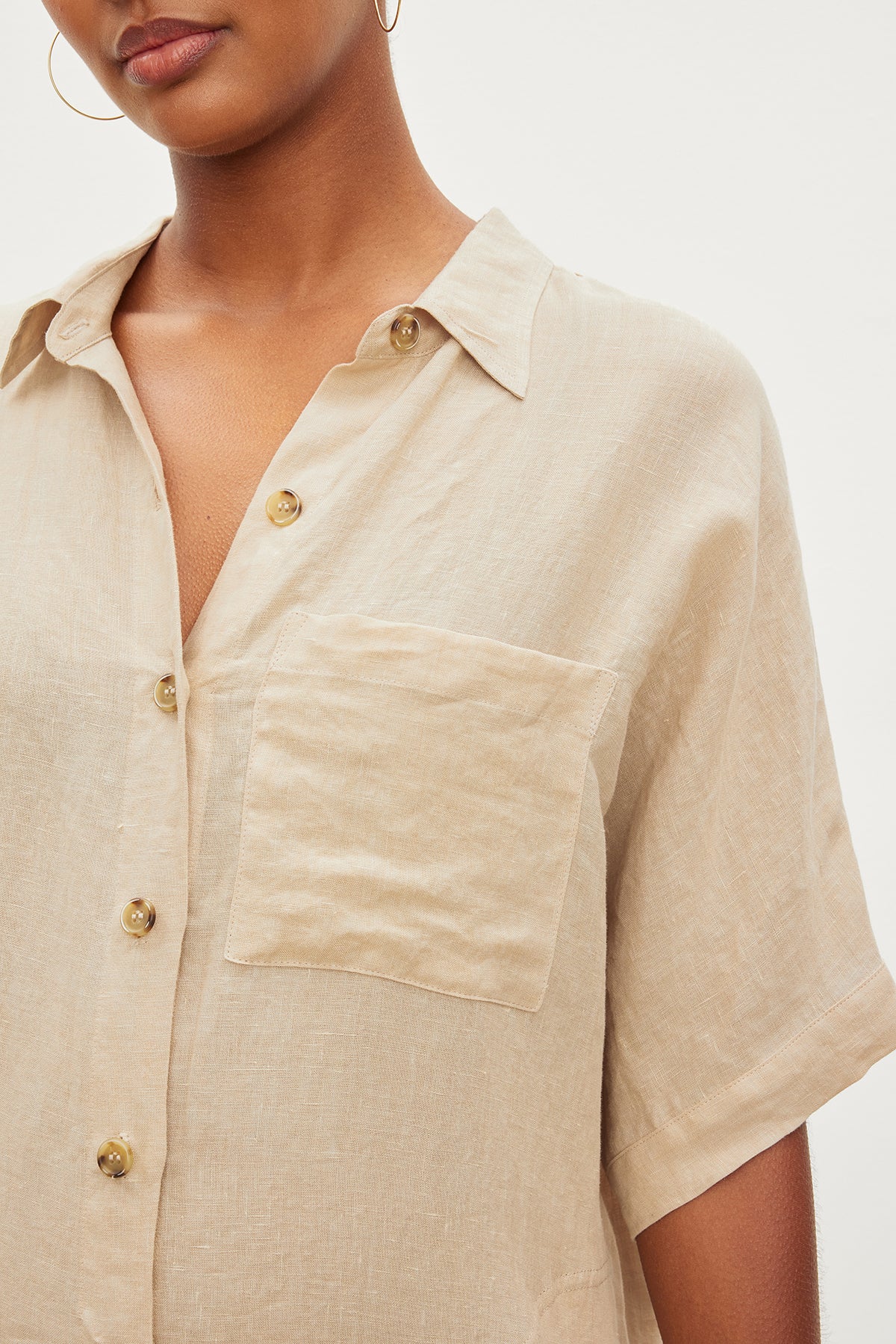   Close-up of a person wearing the Velvet by Graham & Spencer SANDRA LINEN DRESS in light beige, featuring its button-up design and short sleeves. The dress includes a pocket on the left side, and the person accessorizes with a small hoop earring. 