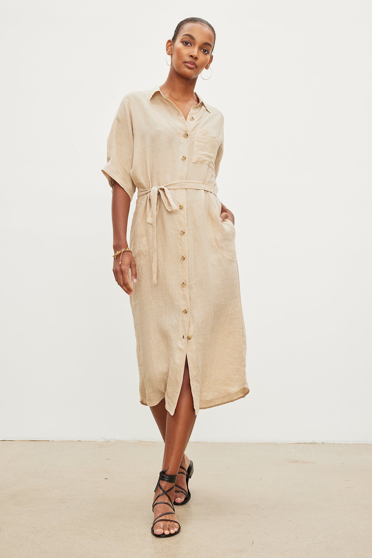 A woman standing against a plain white background wears the SANDRA LINEN DRESS by Velvet by Graham & Spencer. The woven linen midi dress features a button-up design and a detachable belt, paired with black strappy sandals. She has one hand in her pocket and is looking at the camera.-35967717376193