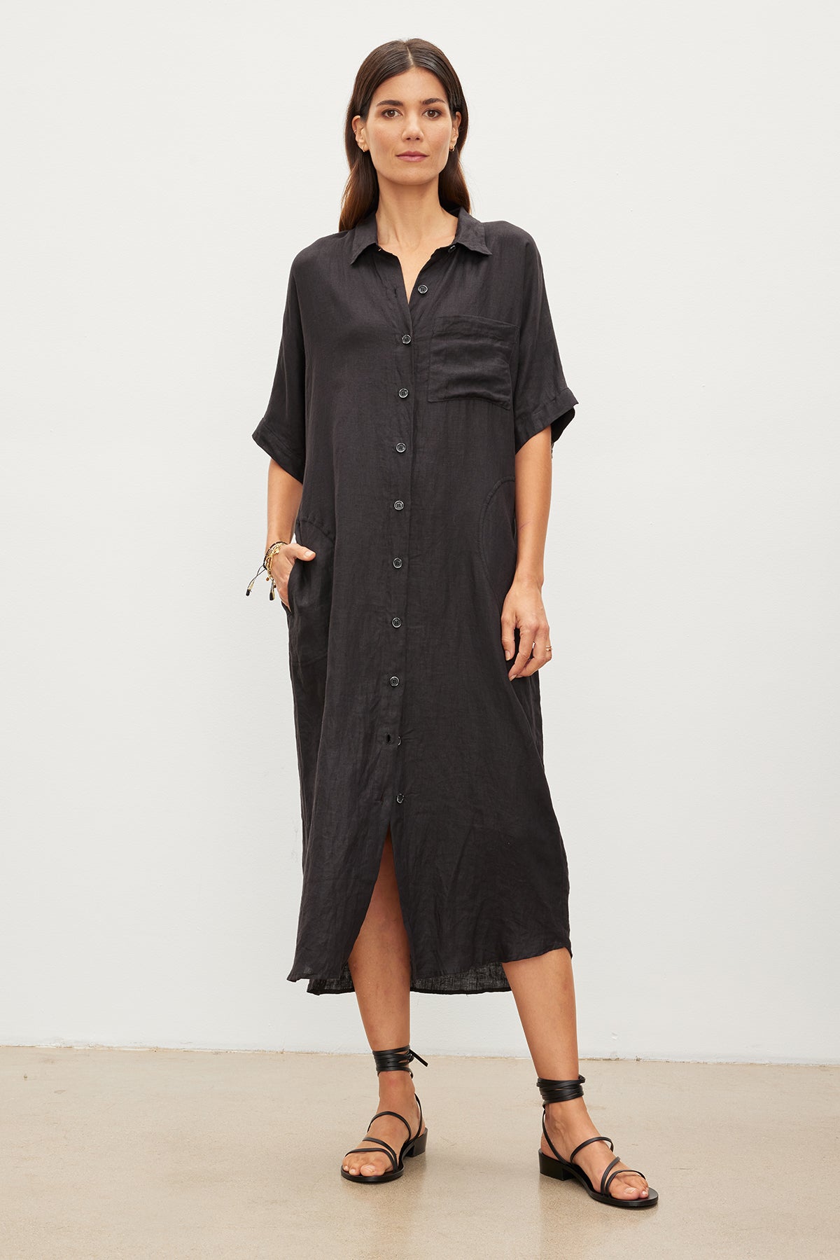   A woman stands against a plain background wearing the SANDRA LINEN DRESS by Velvet by Graham & Spencer, which features a loose-fitting, woven linen fabric, button-up design, and short sleeves. She has one hand in her pocket and wears black strappy sandals. 