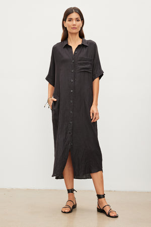 A woman stands against a plain background wearing the SANDRA LINEN DRESS by Velvet by Graham & Spencer, which features a loose-fitting, woven linen fabric, button-up design, and short sleeves. She has one hand in her pocket and wears black strappy sandals.