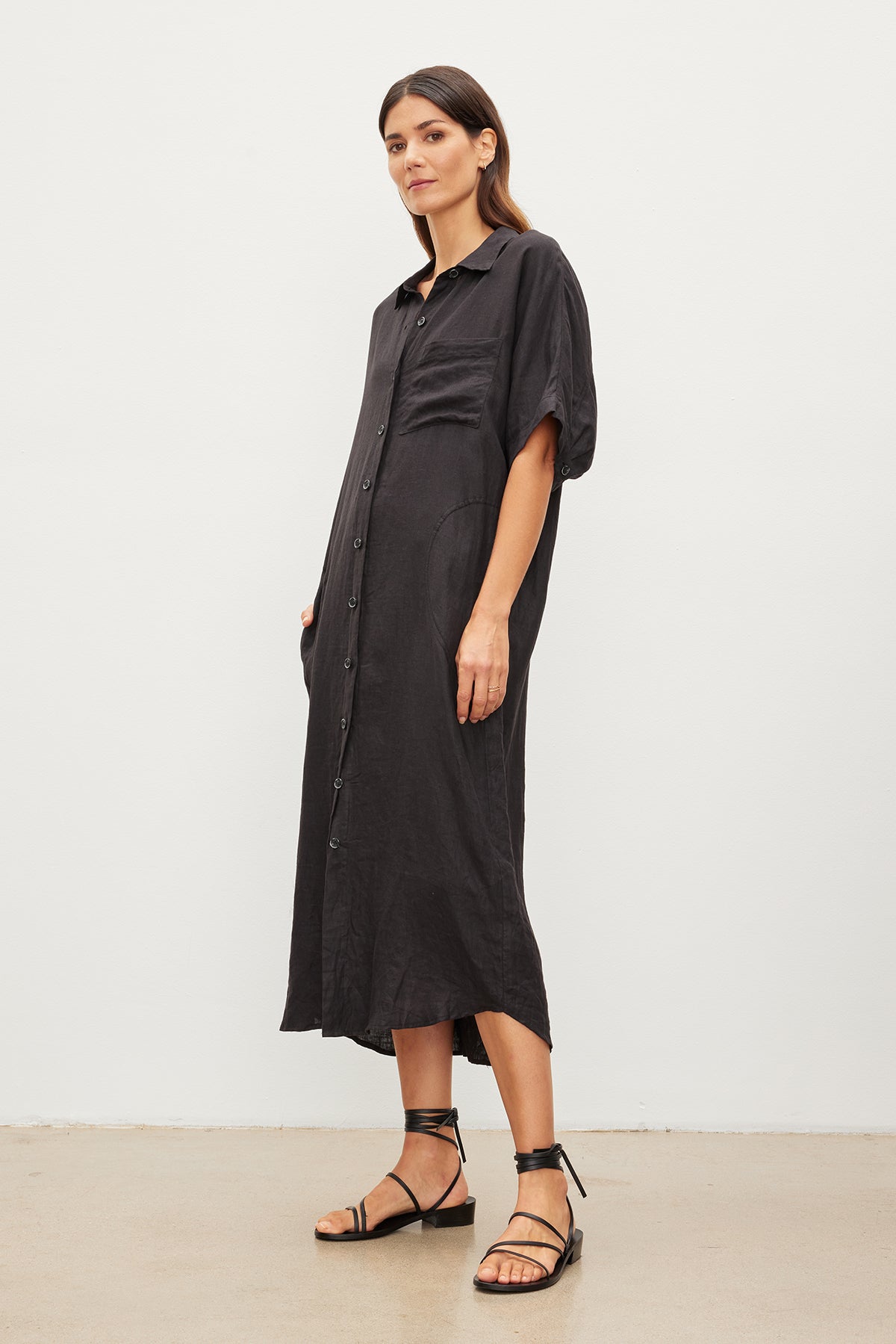 A woman stands against a plain background wearing the SANDRA LINEN DRESS by Velvet by Graham & Spencer, a black, button-up midi dress with short sleeves and a detachable belt, paired with black strappy sandals.-35967717605569