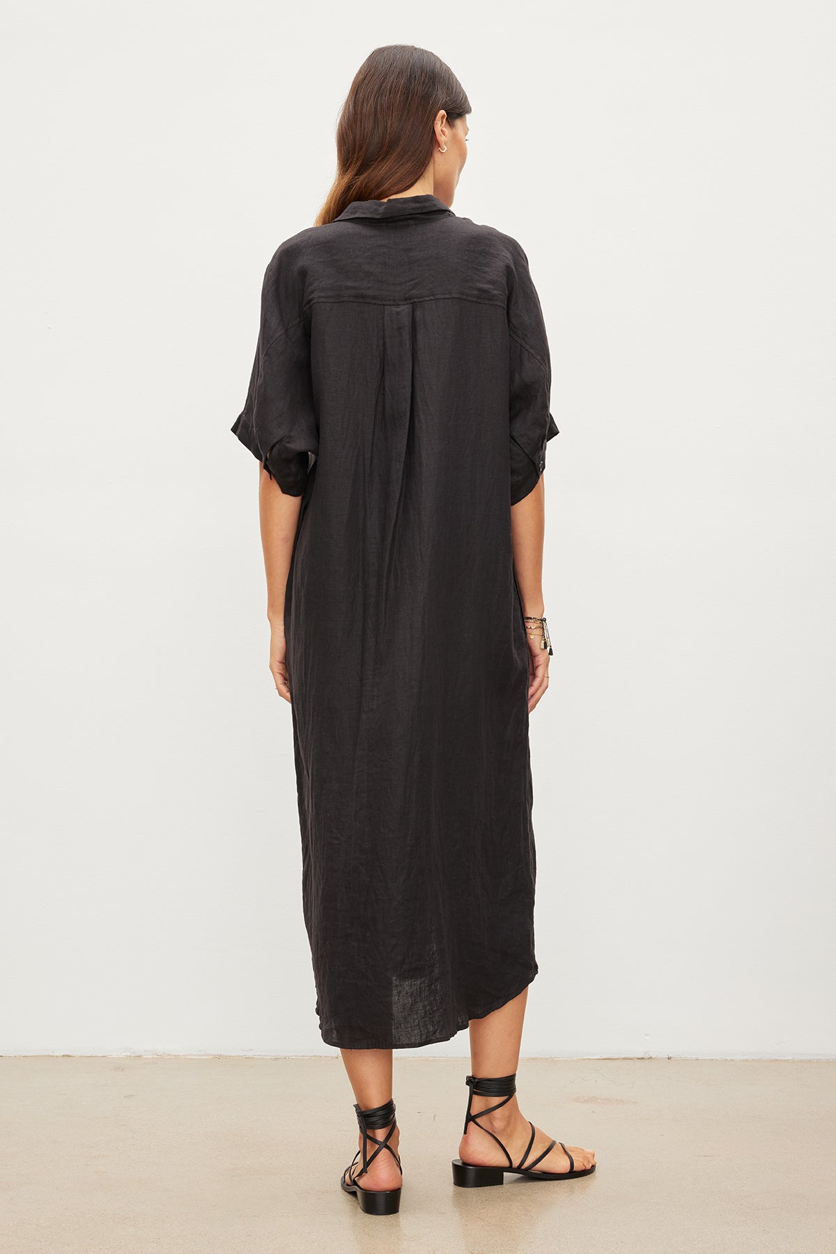 A person with long, dark hair is standing and facing away from the camera, dressed in the SANDRA LINEN DRESS by Velvet by Graham & Spencer. The loose black woven linen dress features a button-up design, paired with black strappy sandals.-35967717638337