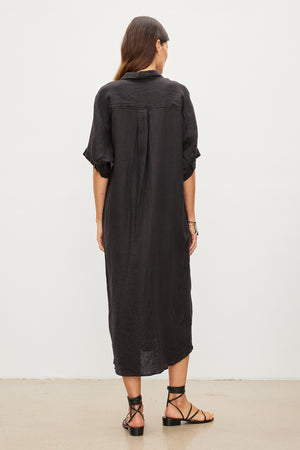 A person with long, dark hair is standing and facing away from the camera, dressed in the SANDRA LINEN DRESS by Velvet by Graham & Spencer. The loose black woven linen dress features a button-up design, paired with black strappy sandals.