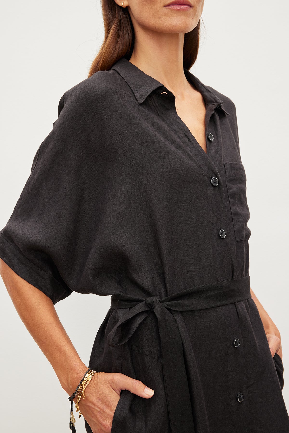   A woman is wearing a black SANDRA LINEN DRESS by Velvet by Graham & Spencer, featuring short sleeves and a detachable belt. She has a minimalist bracelet on her wrist and her hand in her pocket. 