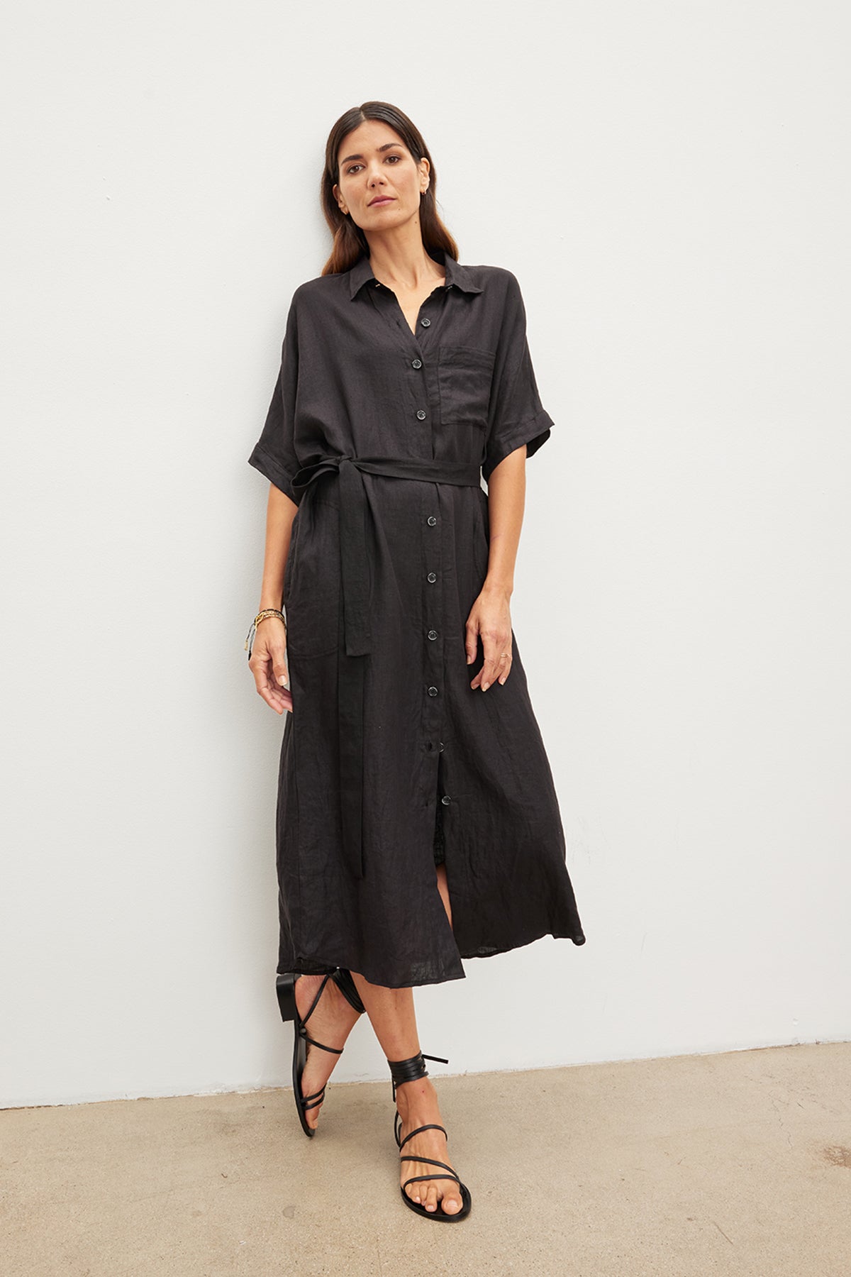   A woman stands against a plain wall, wearing the SANDRA LINEN DRESS by Velvet by Graham & Spencer, featuring a detachable belt, paired with black sandals. She has long dark hair and minimal jewelry. 