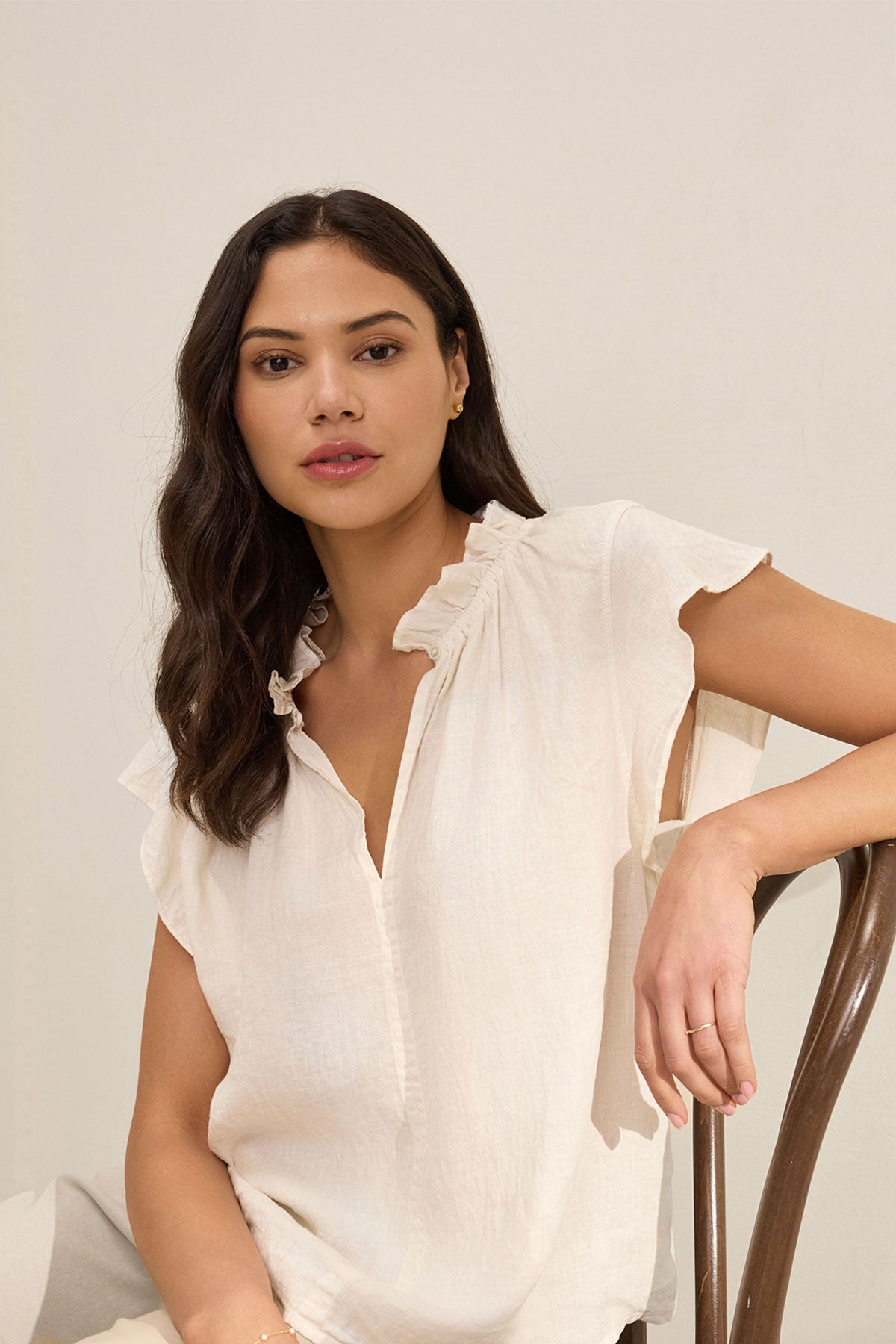 A woman with long dark hair wears the Velvet by Graham & Spencer SEMIRA LINEN TOP, a relaxed fit white blouse with flutter sleeves, while seated on a wooden chair against a neutral background.-39274485022913