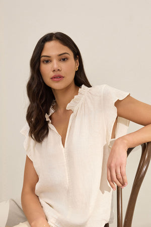 A woman with long dark hair wears the Velvet by Graham & Spencer SEMIRA LINEN TOP, a relaxed fit white blouse with flutter sleeves, while seated on a wooden chair against a neutral background.