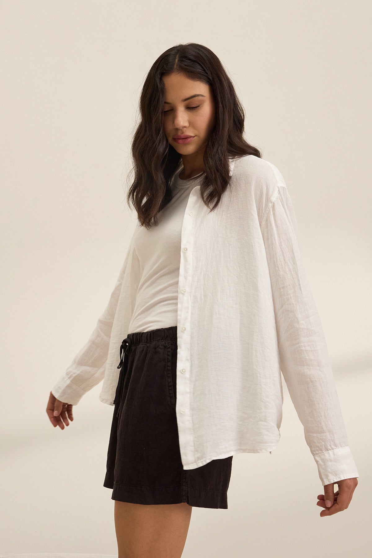 A woman wears Velvet by Graham & Spencer's AMBER LINEN SHORT, paired with a white t-shirt and a lightweight linen button-up shirt, standing against a light plain background, looking down with one arm slightly outstretched.-39261615751361