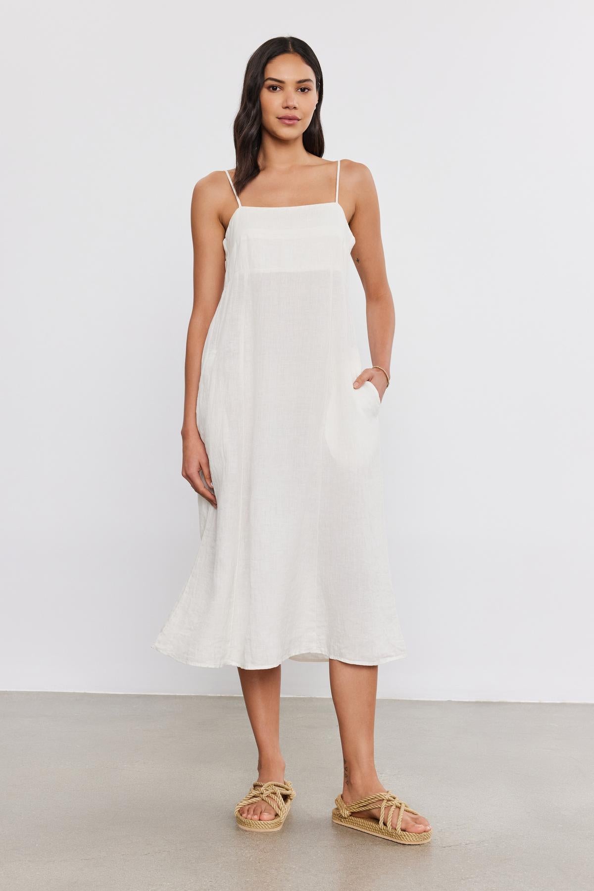 A woman standing in a studio, wearing a white sleeveless Stephie Linen dress by Velvet by Graham & Spencer and straw sandals, and holding a white clutch.-36752944955585