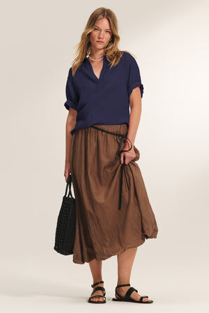 A woman wears the VIVICA LINEN TOP by Velvet by Graham & Spencer, paired with a brown midi skirt, black sandals, and a black woven bag—effortless warm-weather style set against a neutral backdrop.