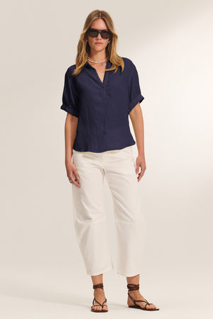 A woman in sunglasses and the Velvet by Graham & Spencer VIVICA LINEN TOP, paired with white linen wide-leg pants, strappy sandals, and a pearl necklace stands against a plain background—perfect for warm-weather dressing.